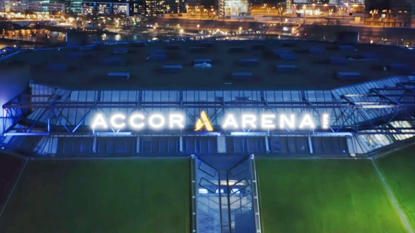 Vista aérea noturna do exterior da Accor Arena, com o letreiro iluminado e as luzes da cidade em segundo plano.