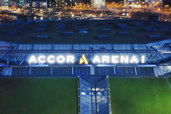 Vista aérea noturna do exterior da Accor Arena, com o letreiro iluminado e as luzes da cidade em segundo plano.