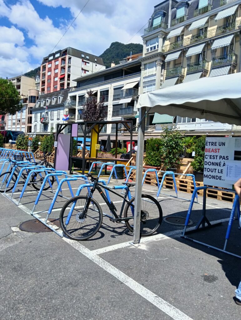 Fila de bicicletas azul-vivo estacionadas num suporte para bicicletas numa praça urbana soalheira, com prédios de apartamentos e uma colina ao fundo