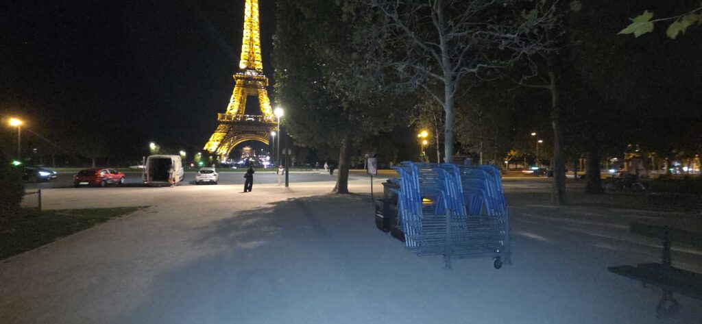 Vista noturna da Torre Eiffel iluminada a amarelo, vista a partir de um parque com cadeiras azuis empilhadas em primeiro plano.